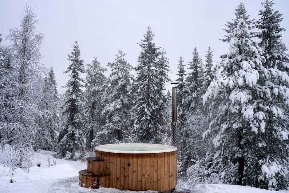A cozy wooden jacuzzi in a snowy winter forest surrounded by pine trees, offering a serene getaway.